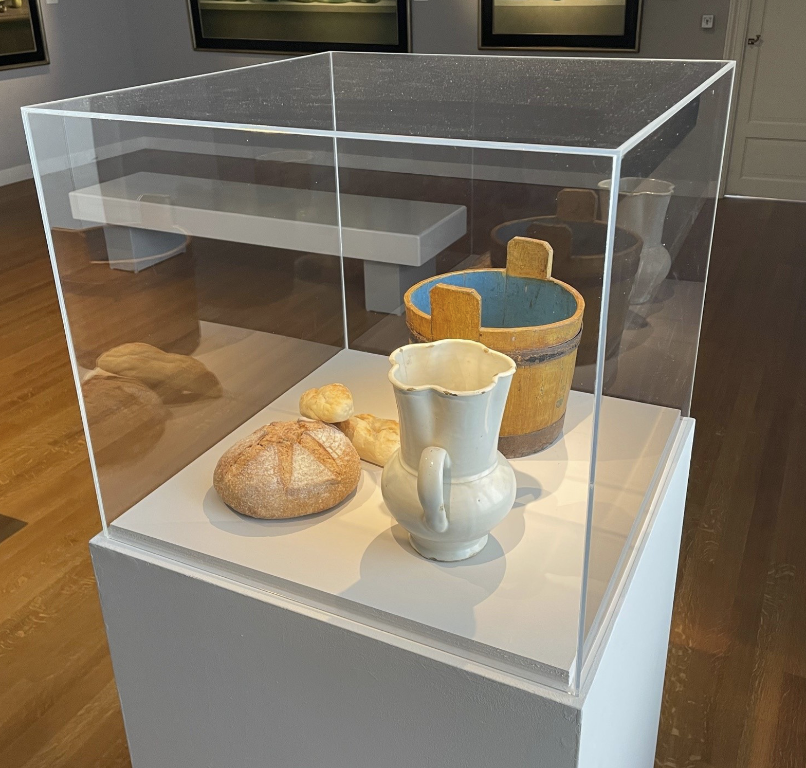 Glazen vitrinekast met brood, witte kan en houten emmer op sokkel in museumruimte met houten vloer