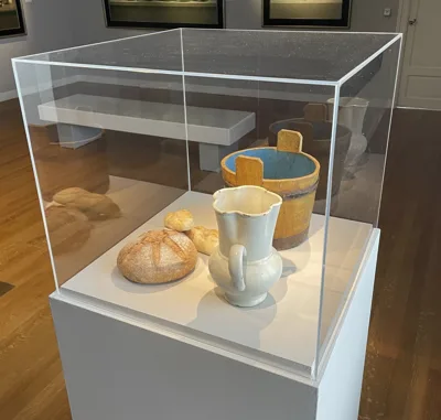 Glazen vitrinekast met brood, witte kan en houten emmer op sokkel in museumruimte met houten vloer
